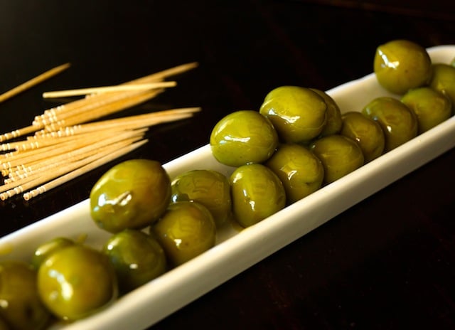 Warm Castelvetrano Olives in a long, diagonal olive dish, with a pile of toothpicks next to it.