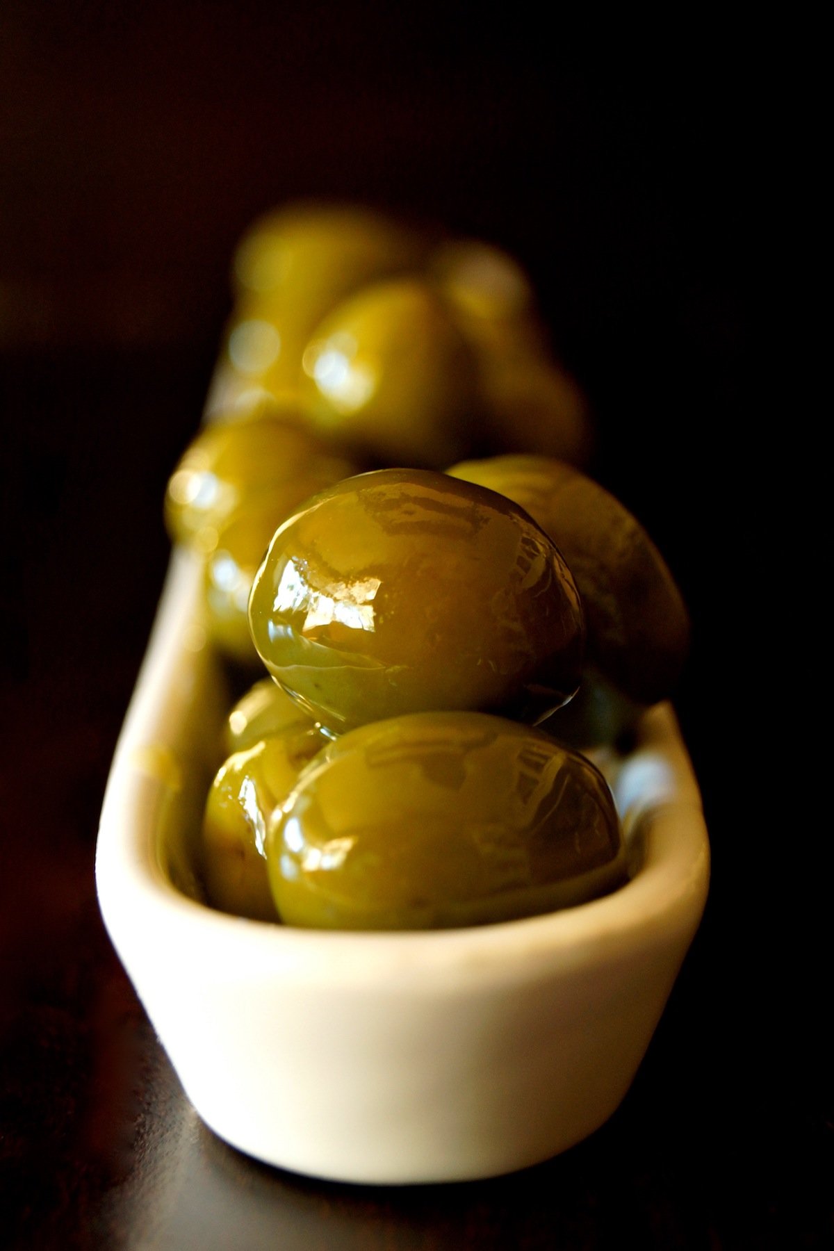 Narrow white olive dish with warm green olives on dark wood.