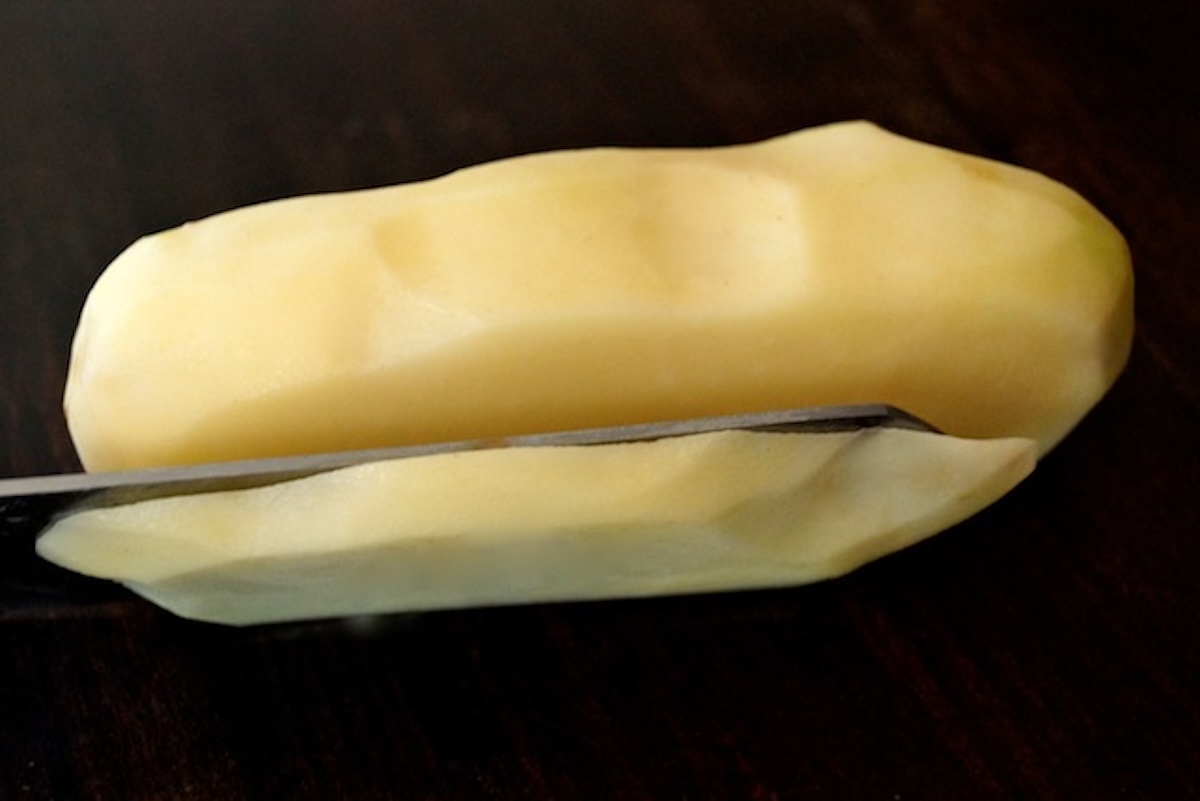 Whole raw and peeled Russet potato with a knife slicing the edge off of it.