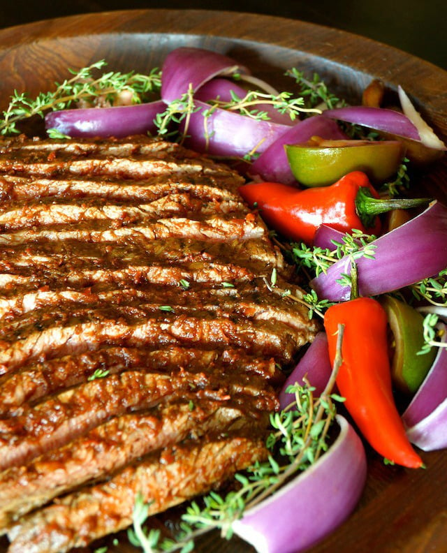 Thinly sliced cooked flank steak on wooden platter with red onion and red peppers.