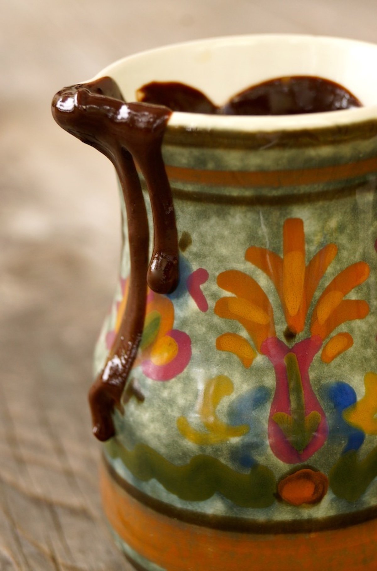 Espresso-Chocolat-Sauce-Recipe Green ceramic pitcher with colorful painted design, dripping from the top with chocolate sauce.
