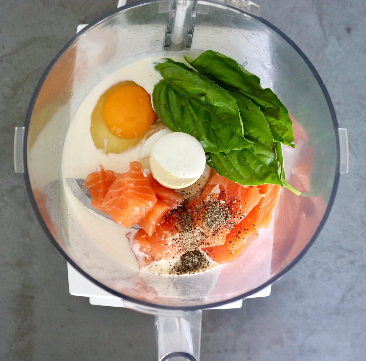 Salmon-and-Cabbage-Recipe Food processor bowl with raw salmon chunks, heavy cream, egg, basil leaves, salt and pepper.