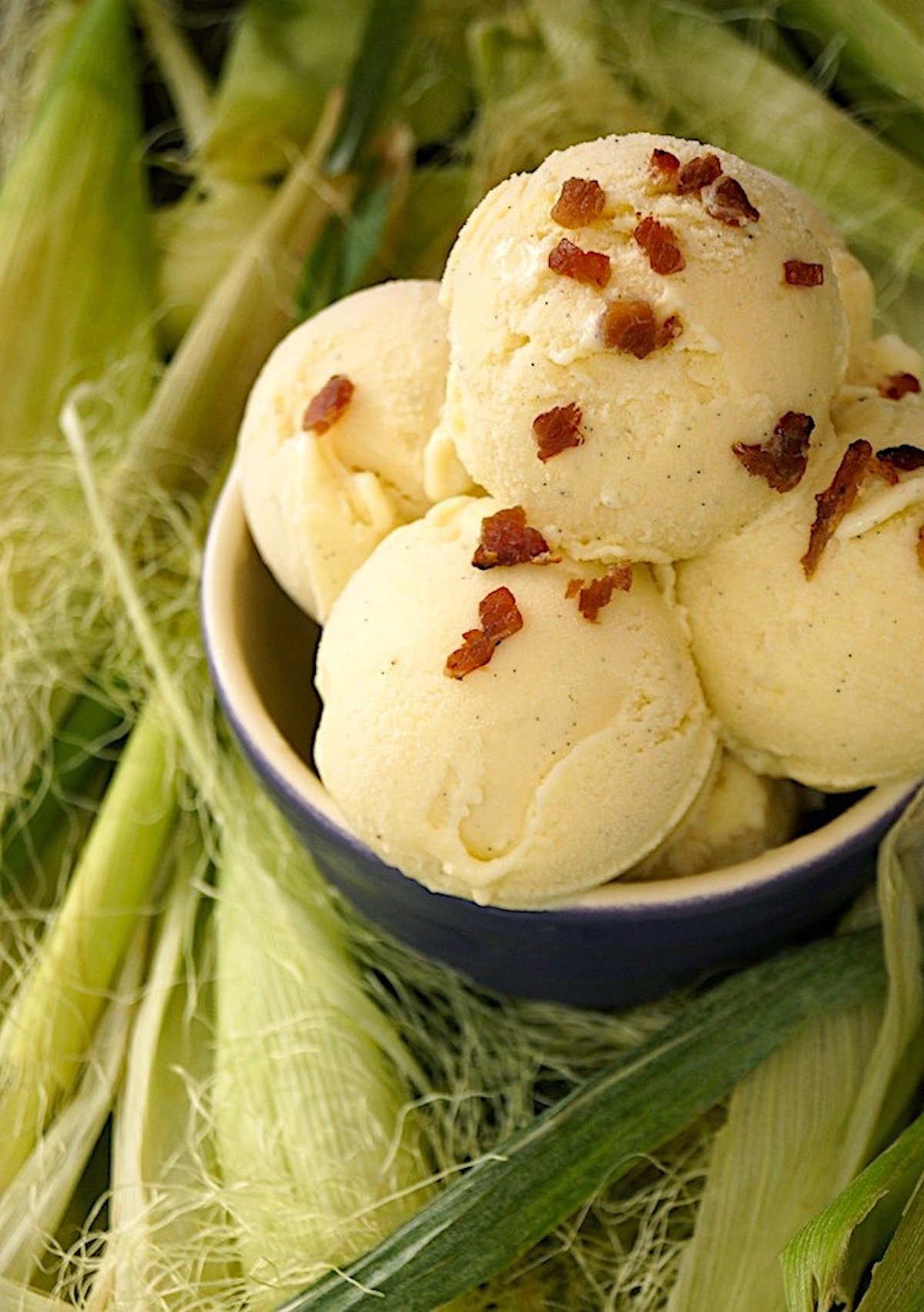 Four scoops of corn ice cream in a purple bowl, surrounded by corn husks.