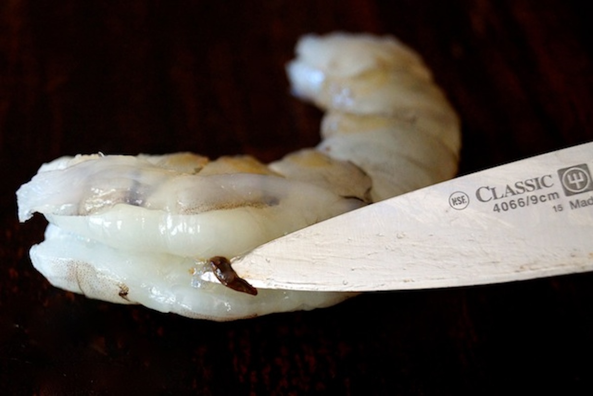 Raw shrimp revealing a bit of the black vein with the tip of a paring knife holding it up.