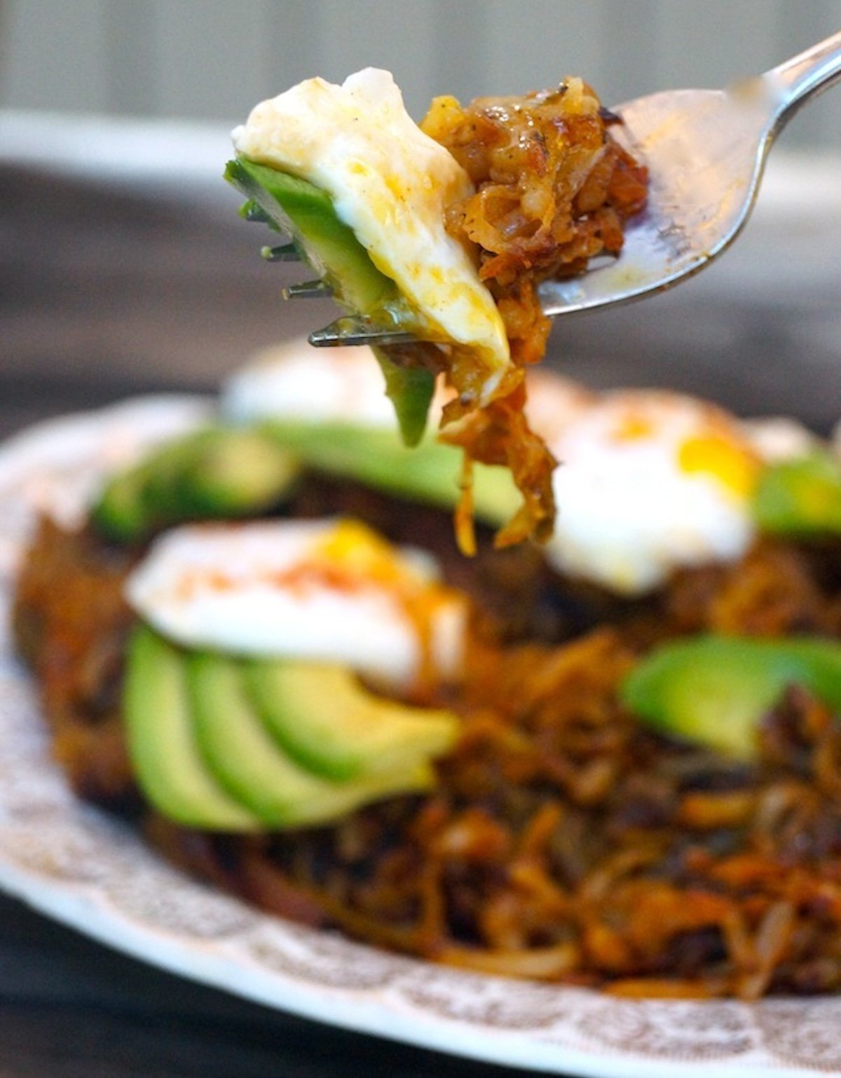 Hash browns on a cream-colored plate with perfect poached eggs and avocado slices on top - with one bite on a fork.