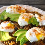 Hash browns with sliced avocado and poached eggs on top, on an oval cream plate.