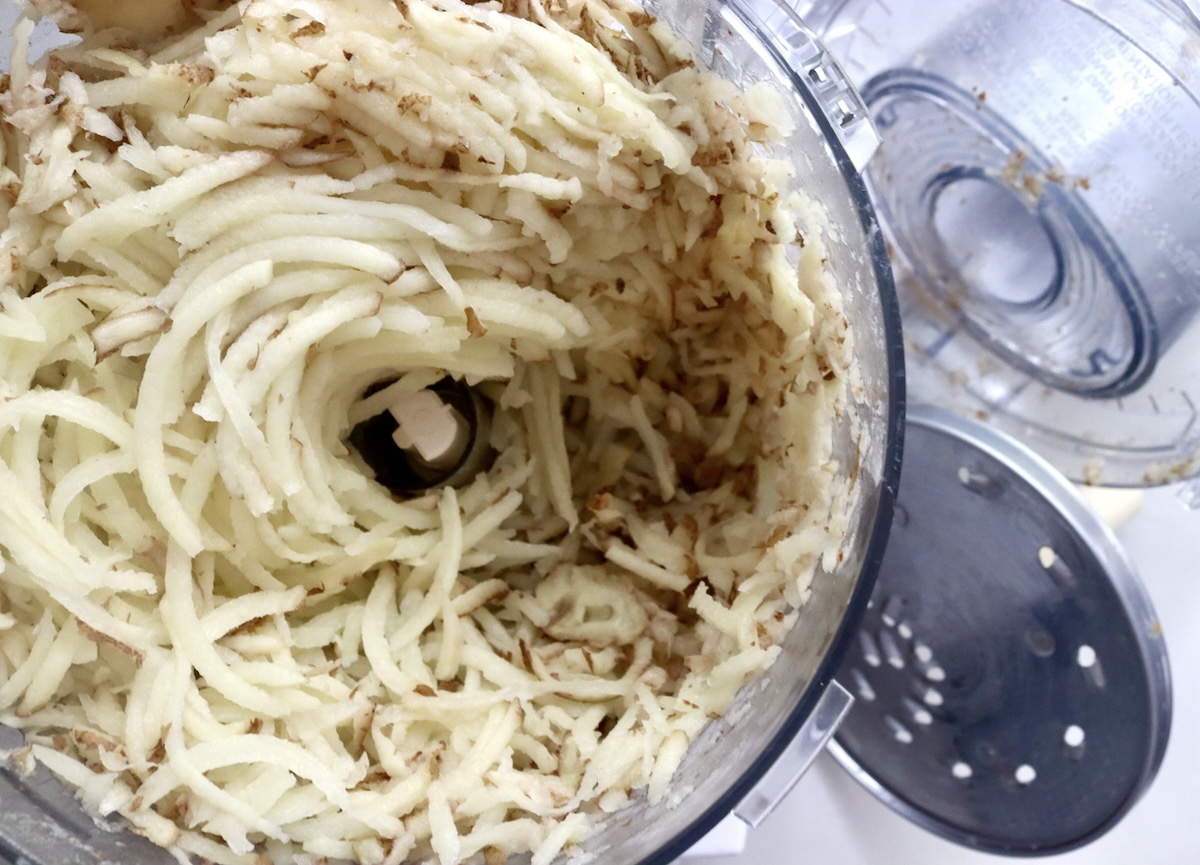 Grated russet potatoes in the bowl of a food processor with the grater attachment next to it.