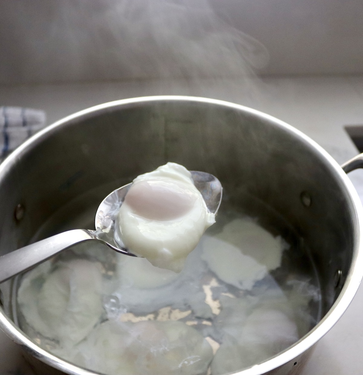 Eggs poaching in the bottom of a large pot, with one being held up in a slotted spoon.