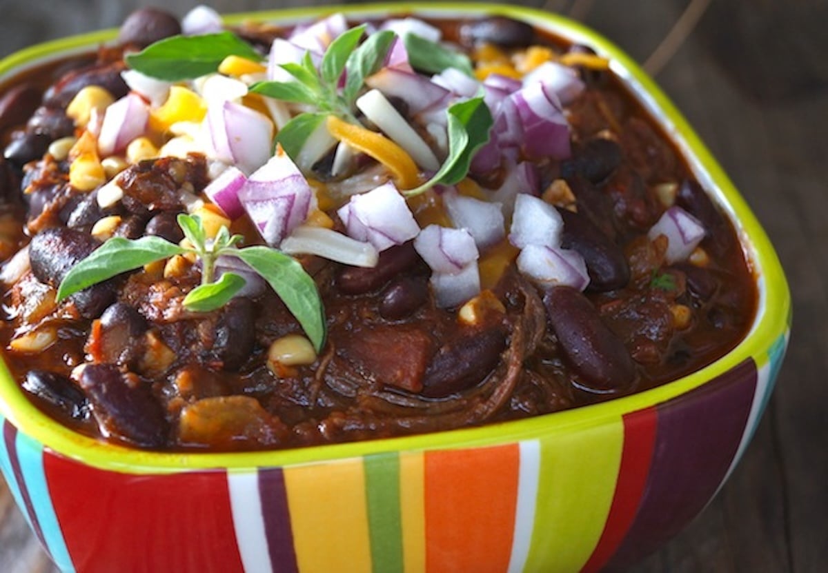 Brisket-Chili-Recipe-with-Bacon-and-Chipotles Rainbow striped bowl filled with chili with red onion and herbs on top,
