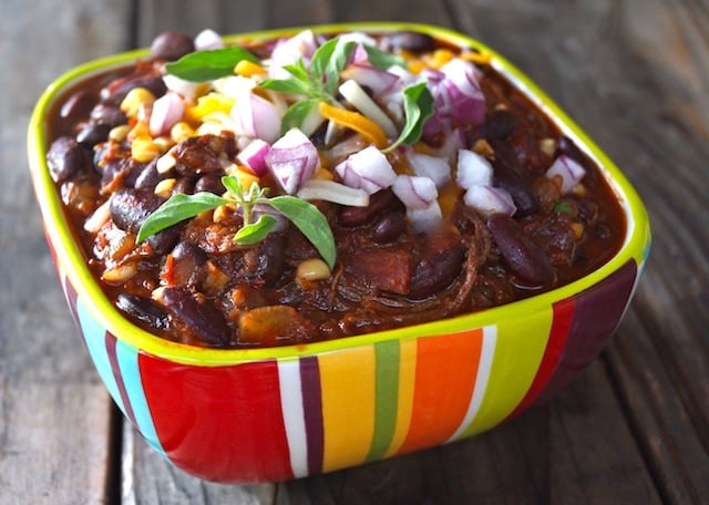 Brisket Chili recipe in a pretty, red, yellow, orange and green striped bowl with fresh oregano, red onion and cheddar cheese.