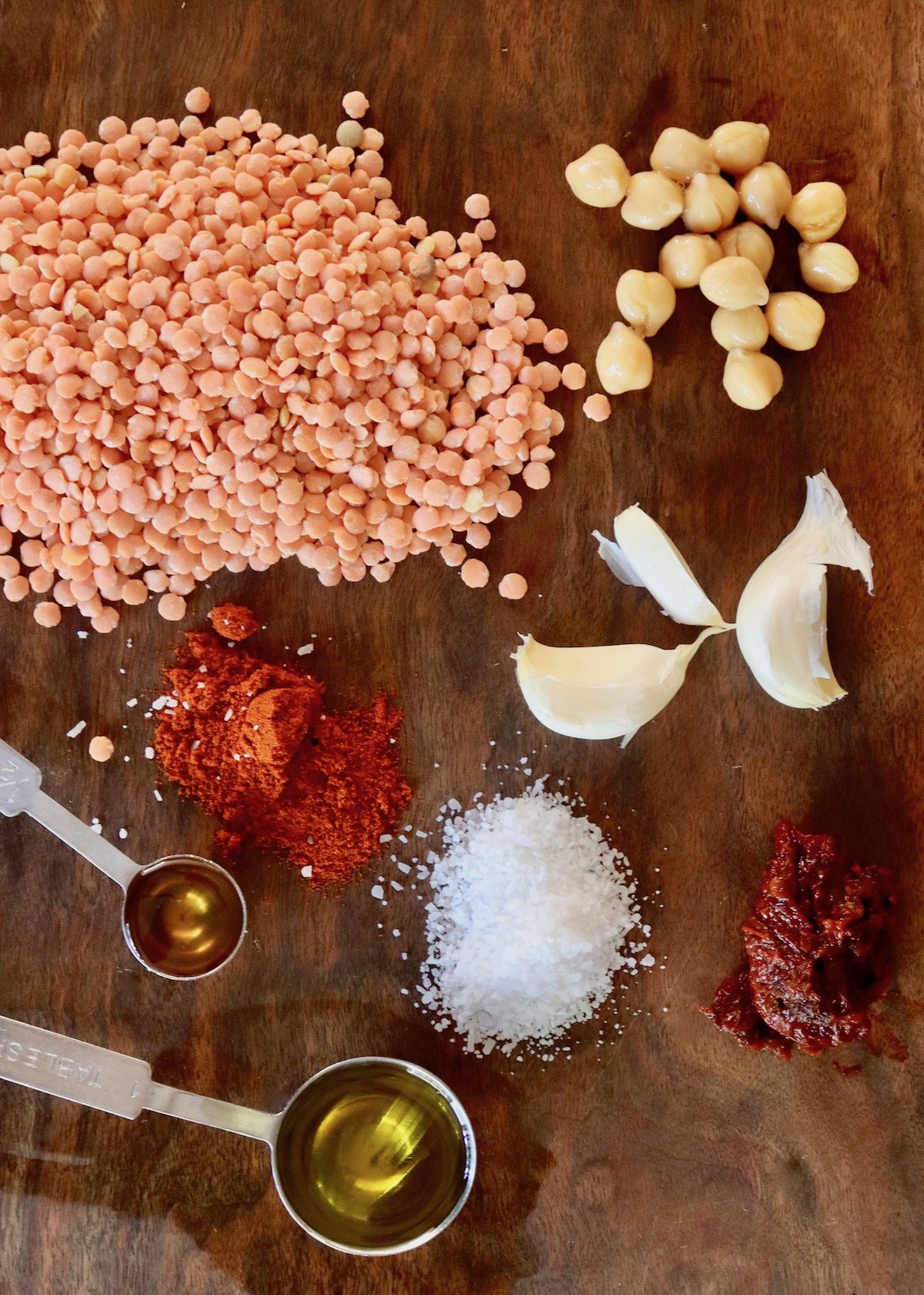 Ingredients for lentil hummus on dark wood cutting board - pile of dry red lentils, garlic cloves, salt, paprika, honey, oil, garbanzo beans and chipotles.
