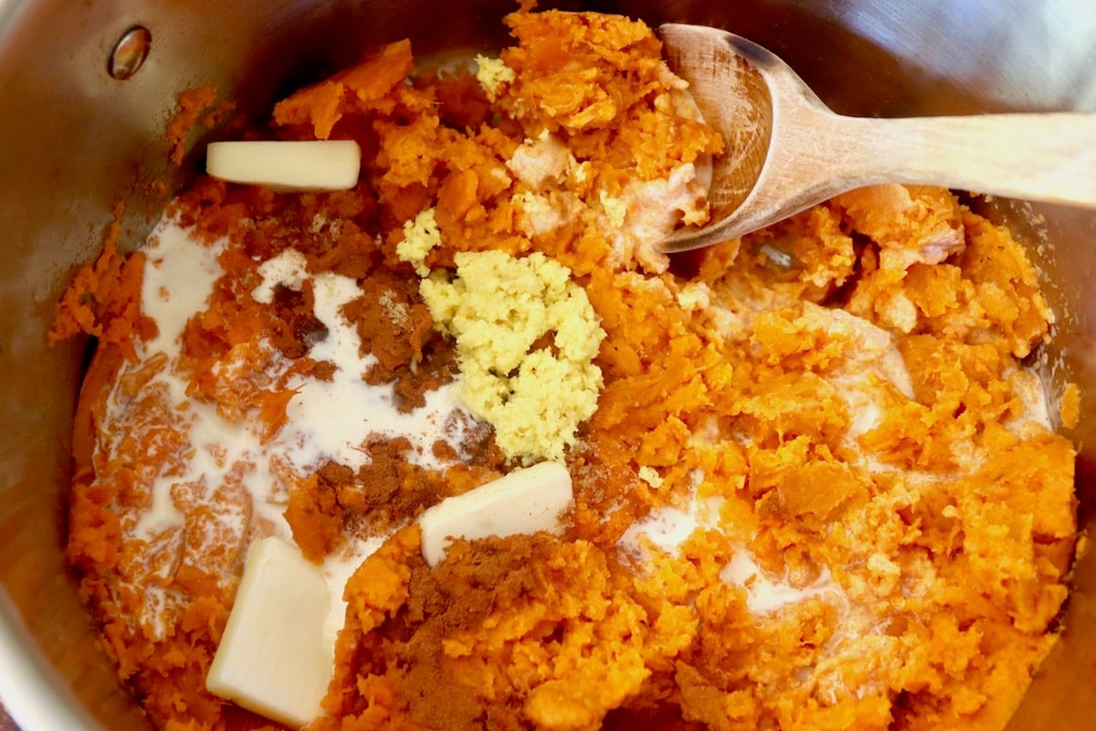 Large bowl filled with mashed sweet potatoes, fresh ginger, butter, cinnamon and cream, with a wooden spoon.