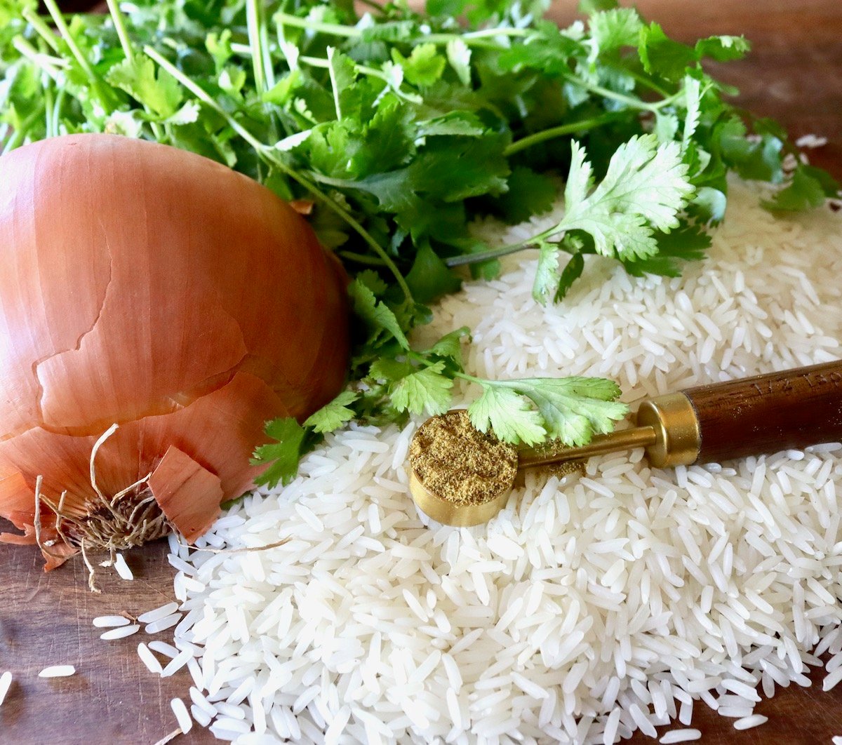 Half of a brown onion, skin side up, with raw white rice, fresh cilantro and a wooden teaspoon with ground cumin.
