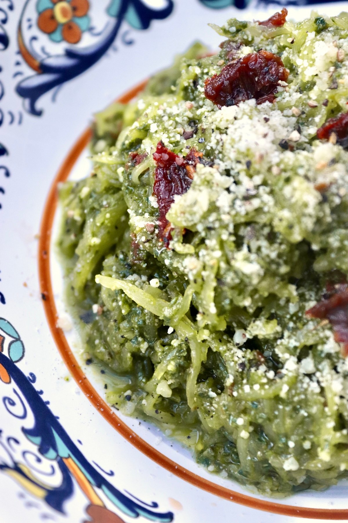 Close up of an Italian-painted ceramic bowl with spaghetti squash tossed with pesto made of spinach and bits of sun-dried tomatoes on top.