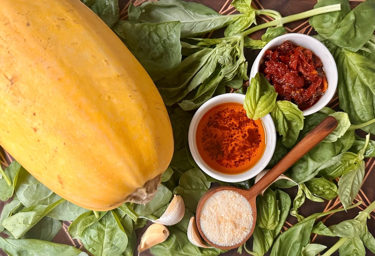 One whole spaghetti squash,  bowls with parmesan cheese, sun-dried tomatoes, sun-dried tomato oil, garlic, spinach and basil on a cutting board.