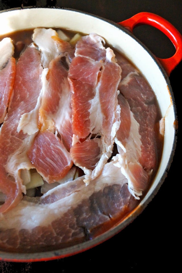 Baked beans in a Dutch oven with a red handle, with a layer of bacon on top.