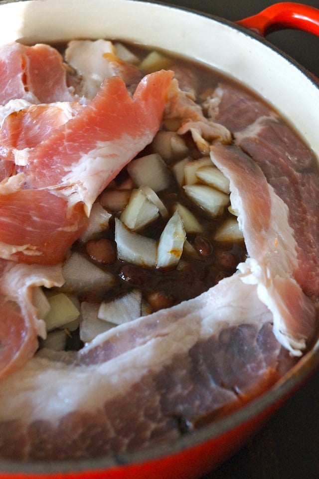 Baked Beans in a Dutch oven with a red handle, with a layer of bacon on top and onions and beans peeking through.