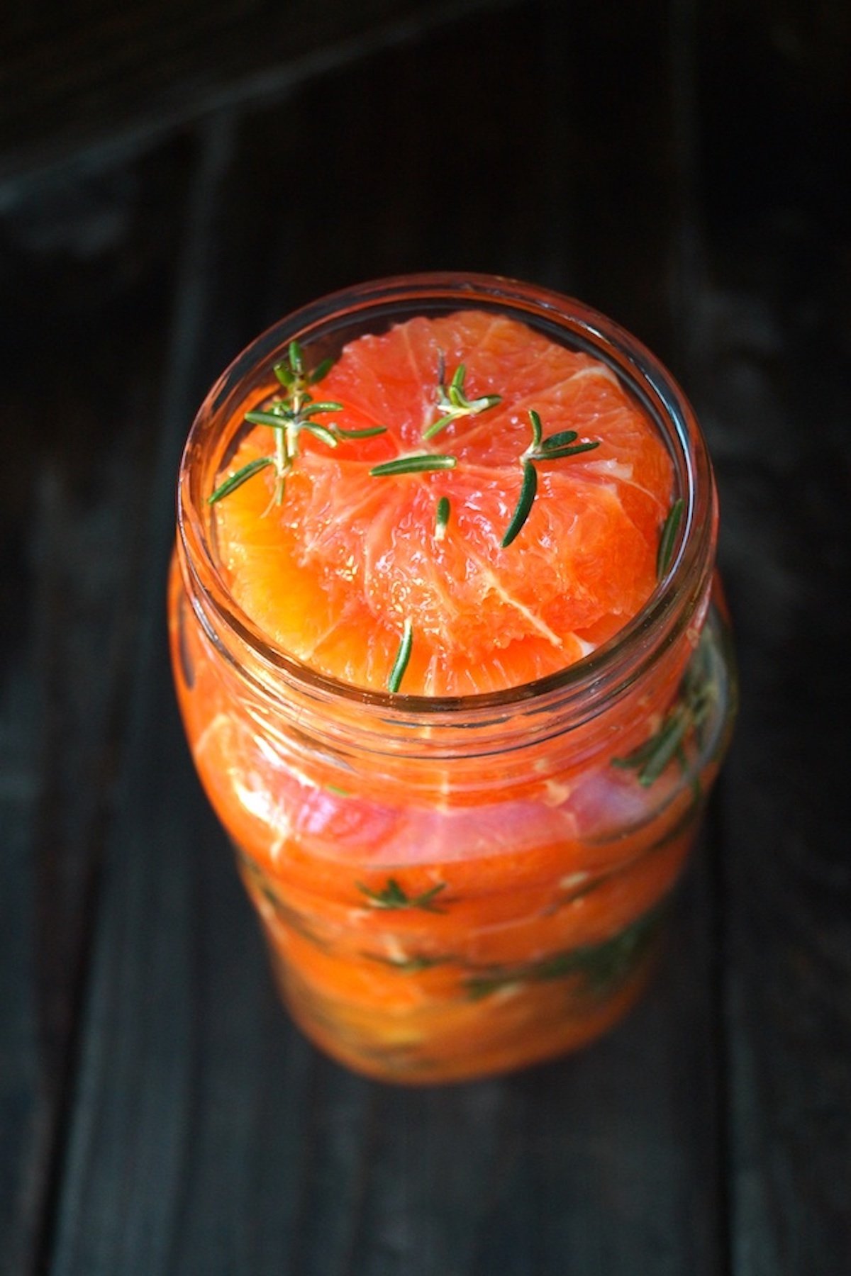Top view of an open jar filled with pink-orange orange slices with rosemary.