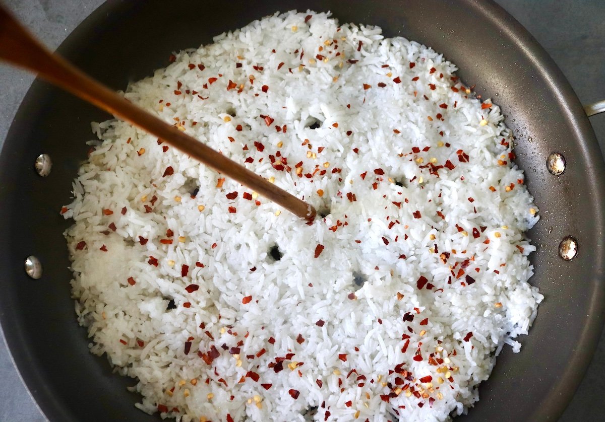Flattened white rice with chili flakes spread on the bottom of a large skillet, and a long wooden stick making holes in it.
