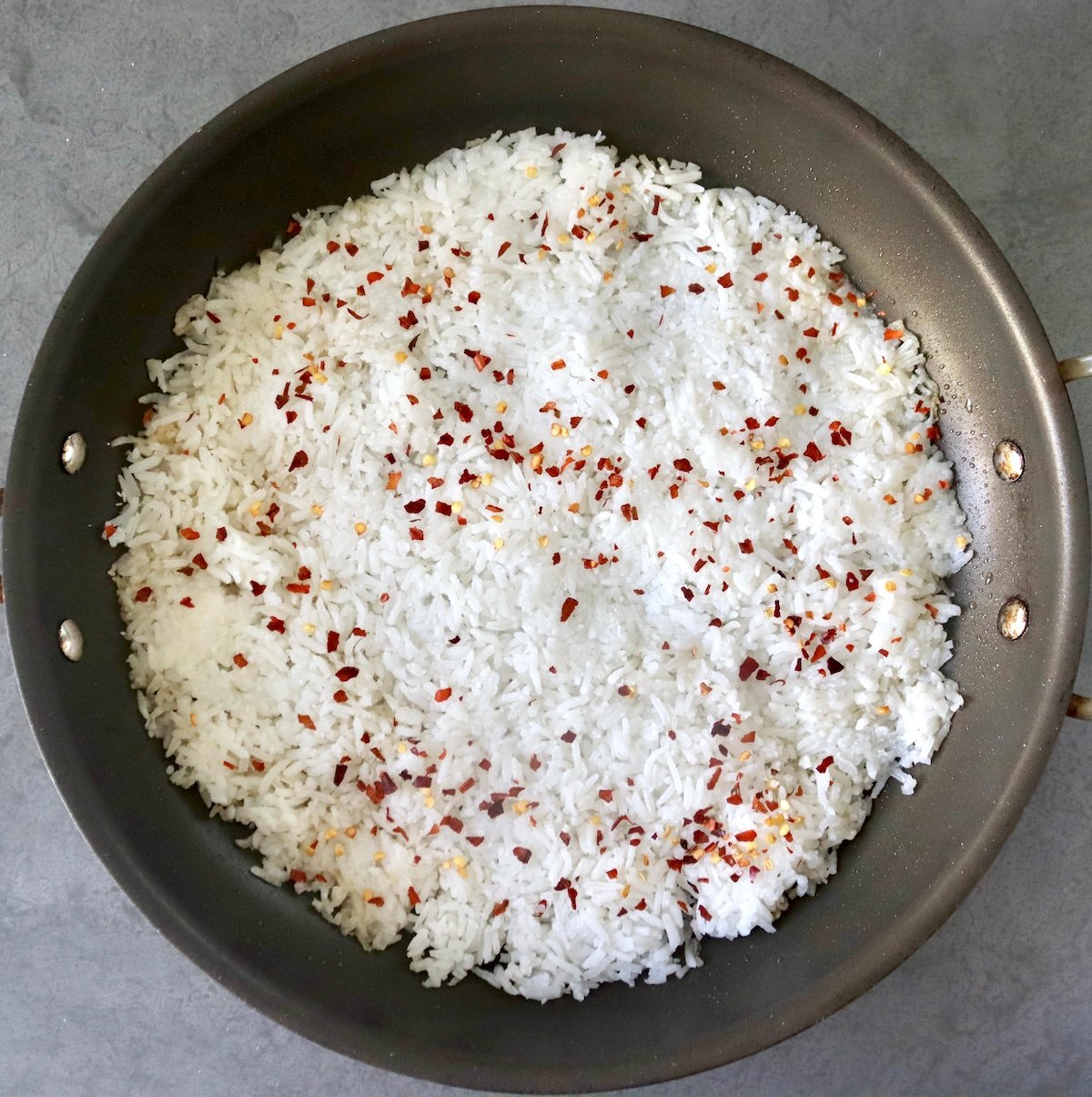 Flattened white rice with chili flakes spread on the bottom of a large skillet.