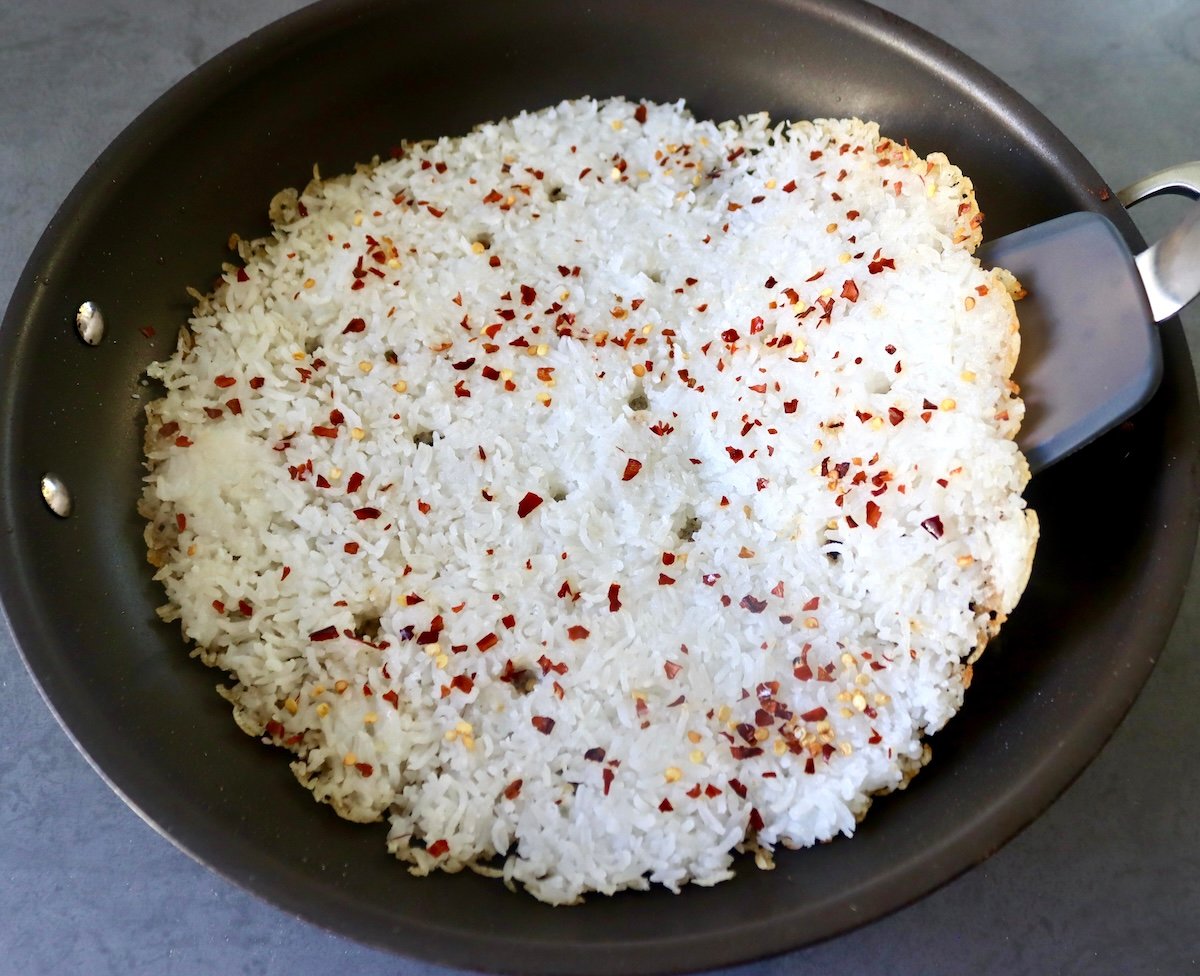 Flattened white rice with chili flakes spread on the bottom of a large skillet, and being lifted with a black rubber spatula.