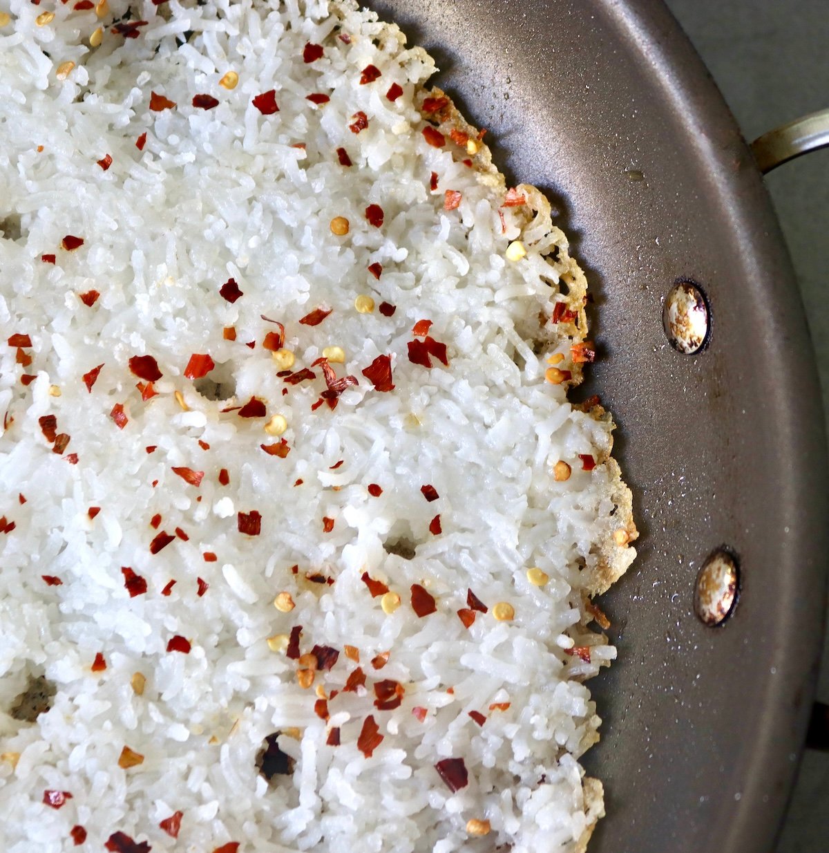 Close up of flattened rice in a skillet with chili flakes, with a golden crispy edge.