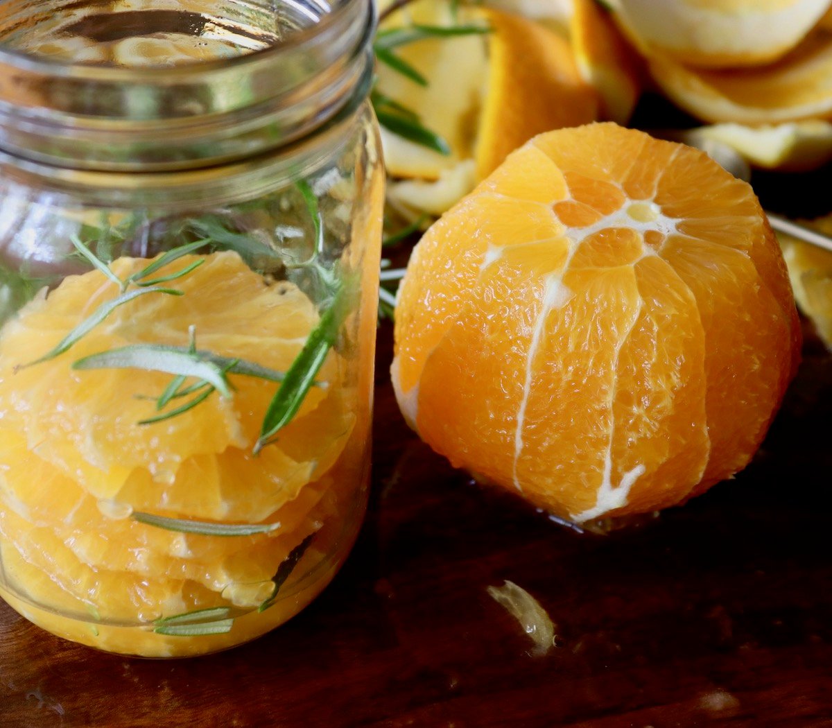 Jar about half filled with orange slices and rosemary with peeled orange next to it.