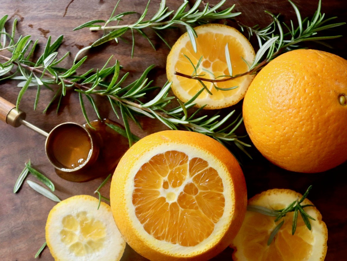 Two rosemary sprigs, tablespoon full of honey and cut oranges, on cutting board.