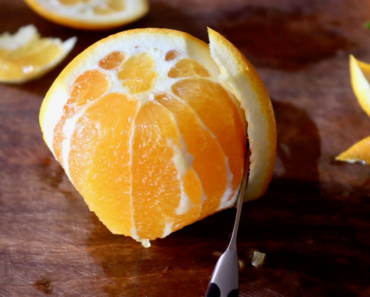 One orange with the skin being sliced off with a paring knife, on cutting board.