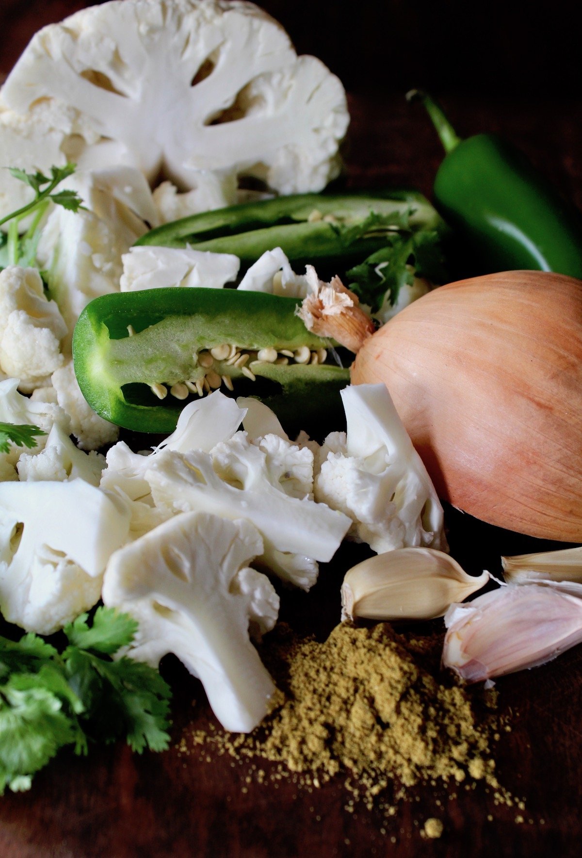 Garlic, cauliflower, jalapeno, ground cumin and half onion of cutting board.