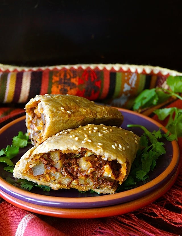 One Potato Chorizo Hand Pie cut in half on a ceramic, purple plate with fresh cilantro and a Mexican-style cloth.