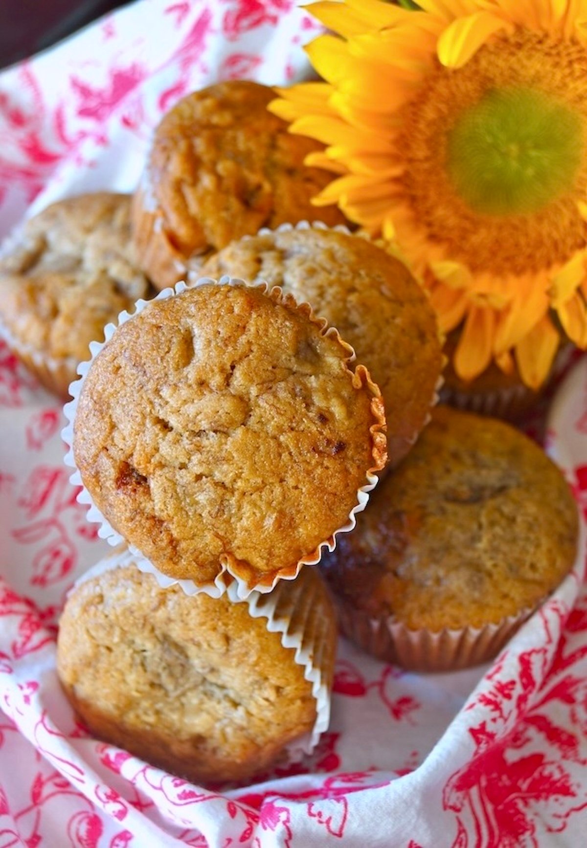 Pink and white floral cloth and 1 big yellow sunflower, with several golden honey muffins on top.