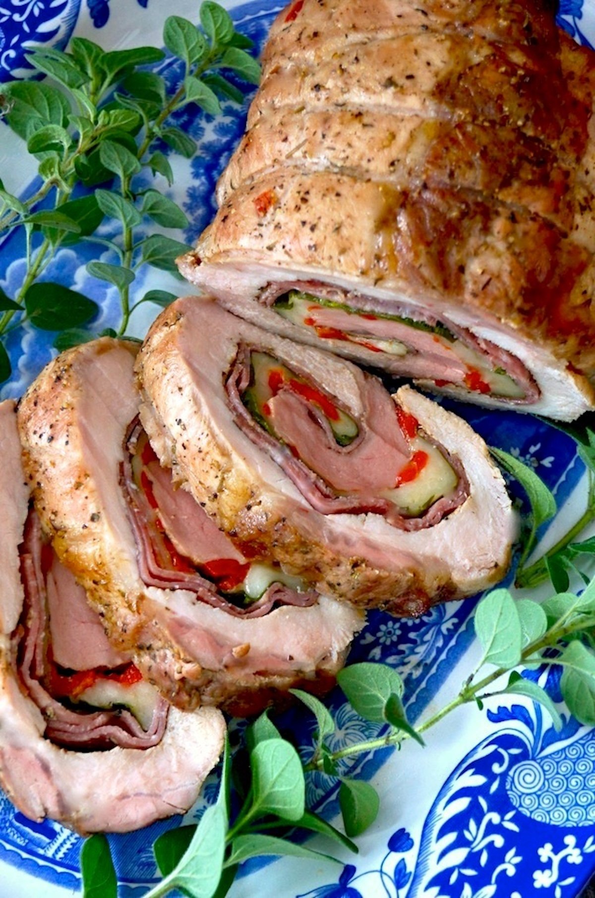 Sliced and roasted stuffed pork loin with salami, provolone and red pepper, on a pretty blue and white plate with fresh oregano sprigs.