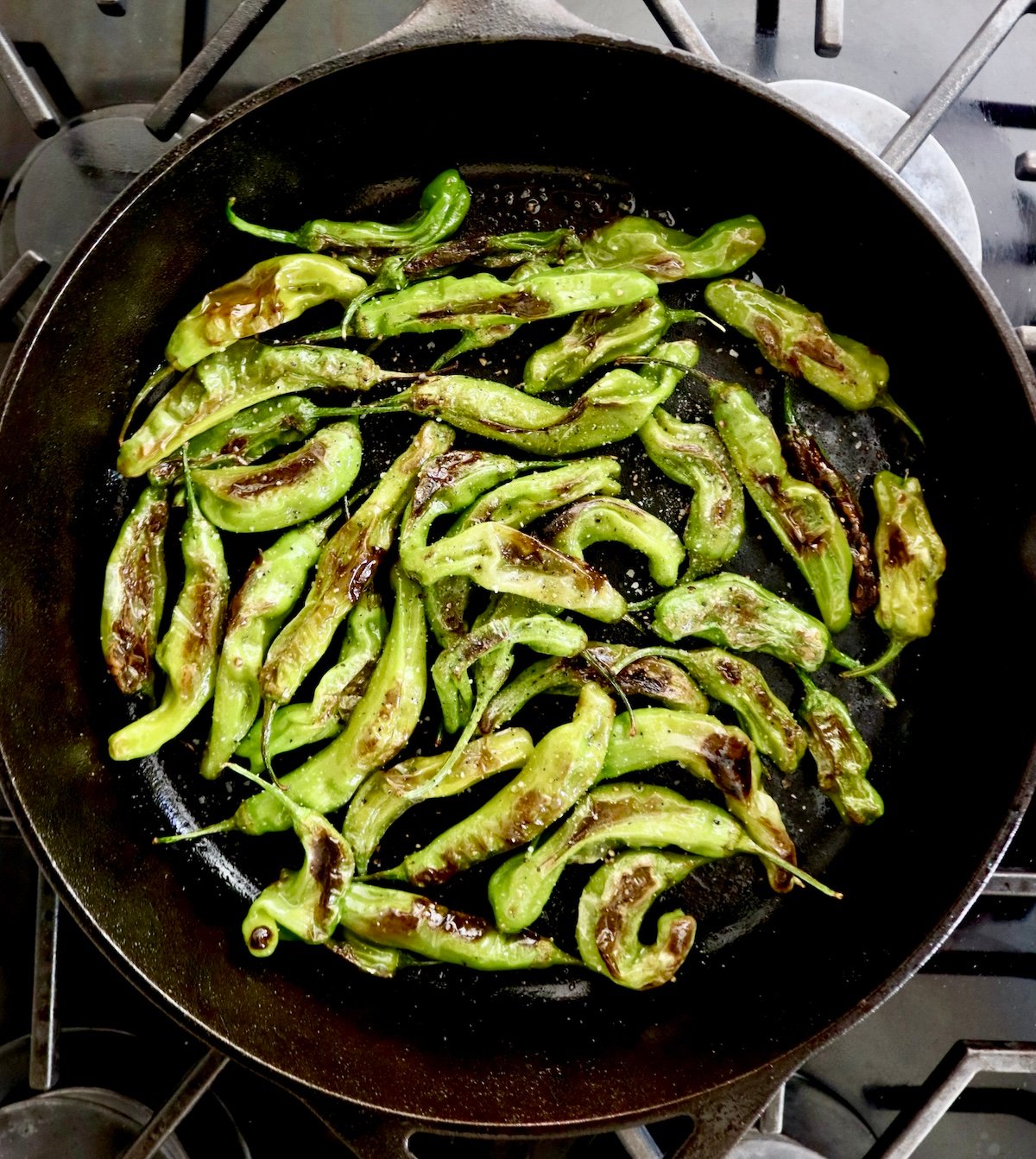 Charred and blistered bright green shishito peppers in a cast iron skillet sprinkled with salt and pepper.
