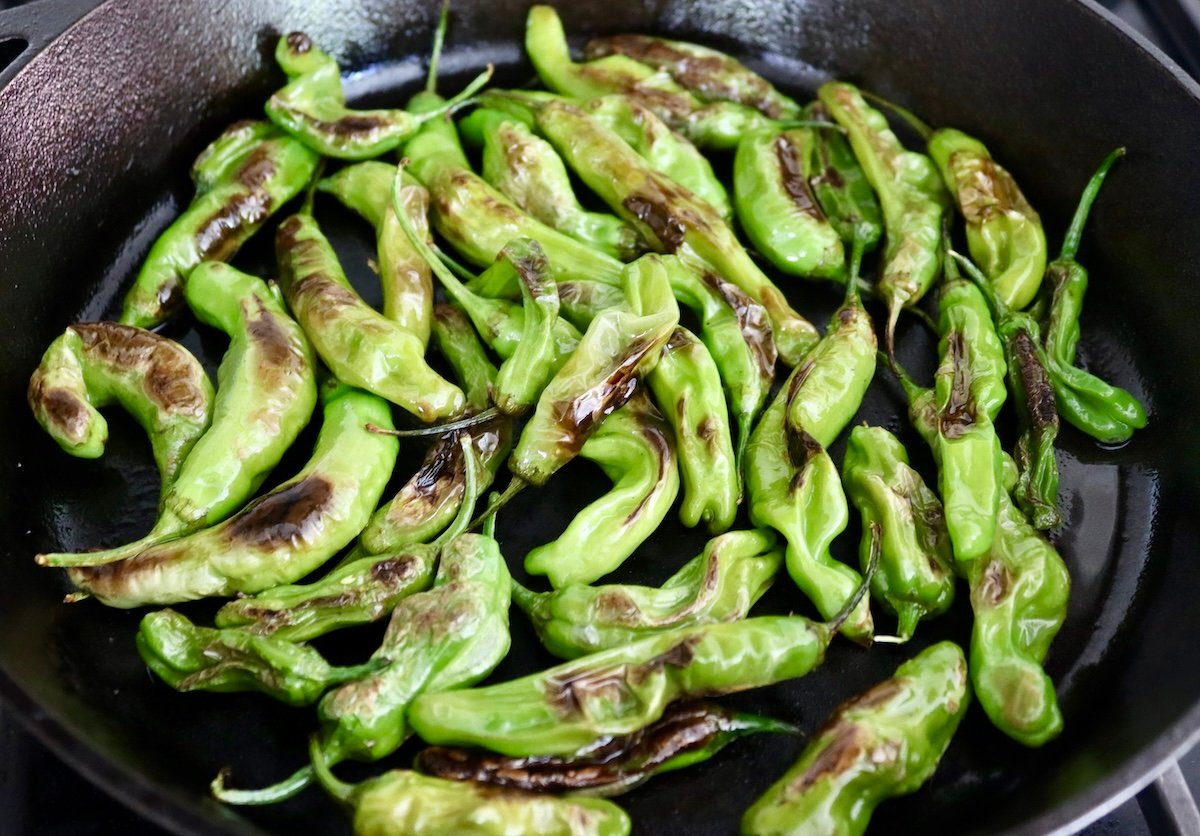 Charred and blistered bright green shishito peppers in a cast iron skillet.