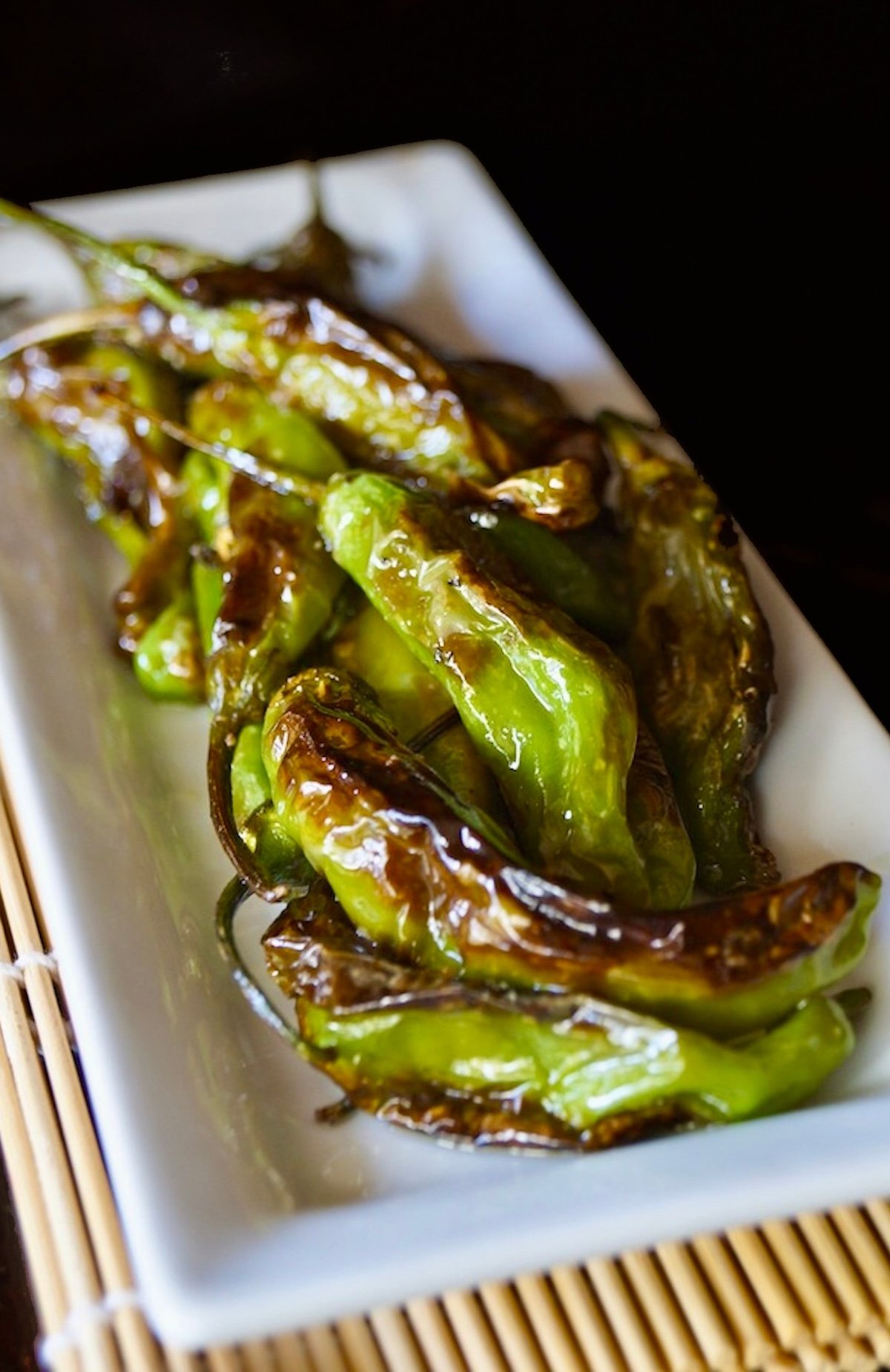 Blistered-Shishito-Peppers White rectangular plate on a bamboo sushi mat with charred shishito peppers.