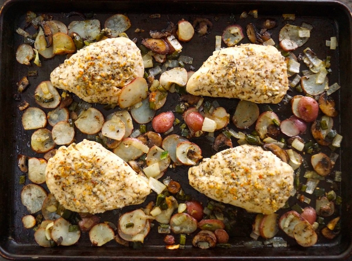 Four baked Parmesan coated chicken breasts on a sheet pan with vegetables. 