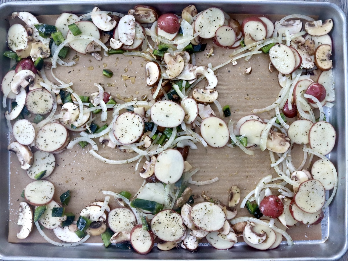 Sheet pan with parchment paper and vegetable mix of sliced potatoes and mushrooms spread out with 4 spaces for chicken.