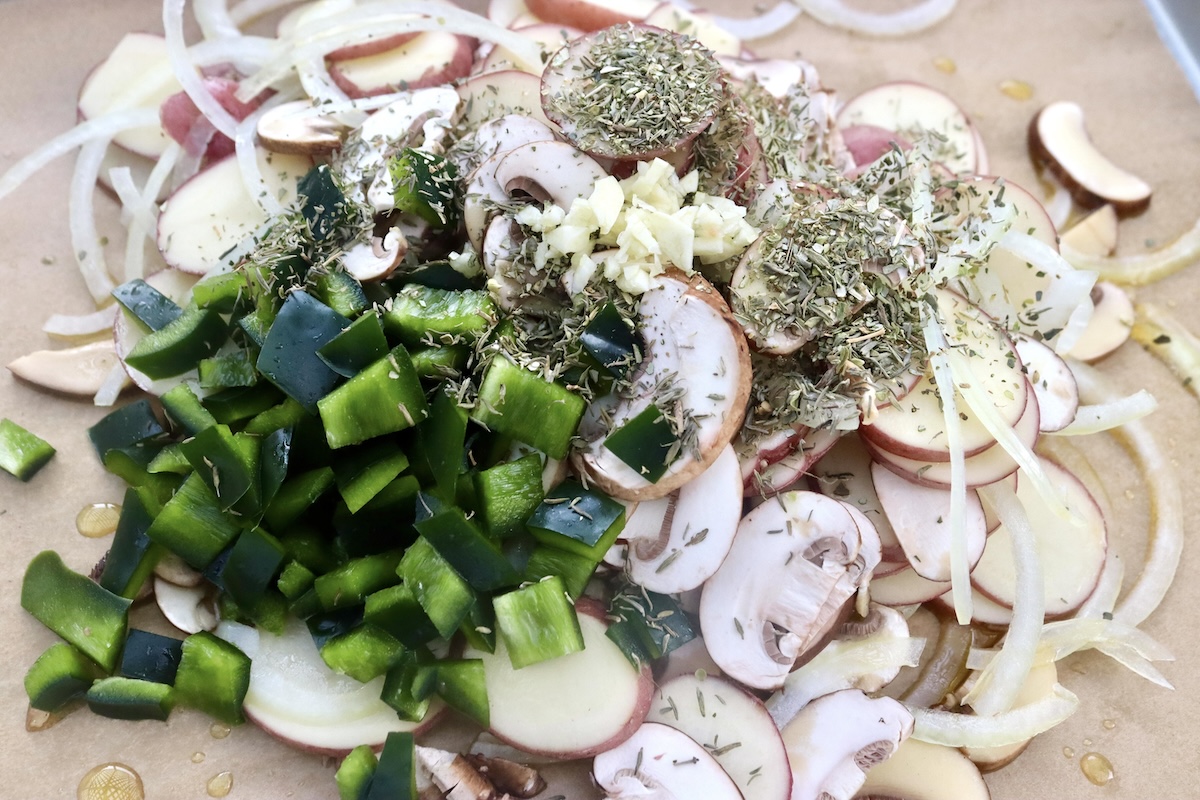 Pile of thinly sliced red potatoes, dried herbs, minced garlic, sliced mushrooms and diced poblano peppers, on a sheet pan.