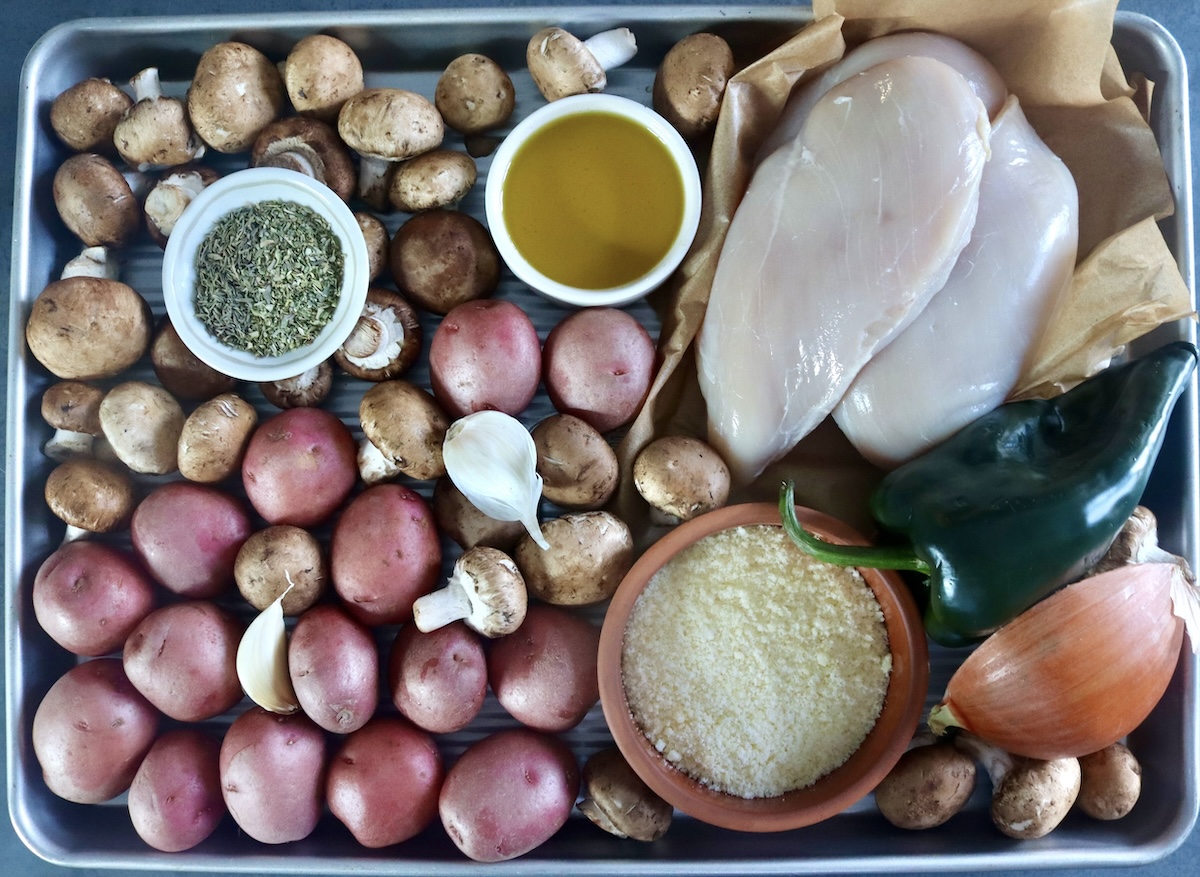Sheet pan filled with small red potatoes, raw chicken, whole crimini mushrooms, a poblano pepper, garlic cloves, and parmesan cheese, herbs and olive oil in small bowls. 