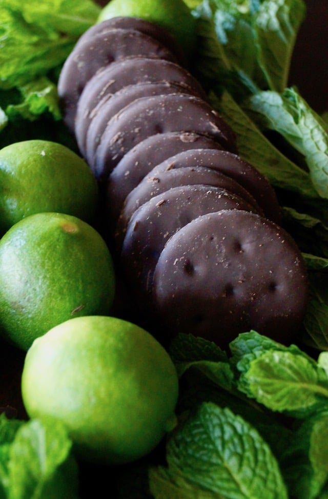 Three limes and a row of Thin Mint Girl Scout cookies surrounded by fresh mint leaves.