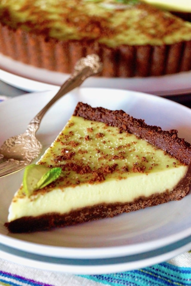 One slice of Thin Mint Key Lime Tart with whole tart behind it, on a white plate with a silver fork.