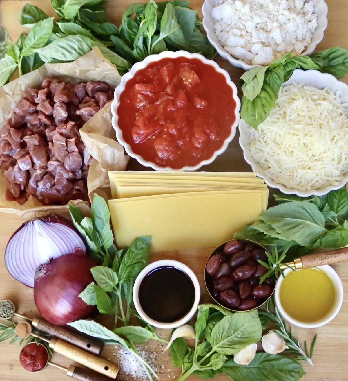 Lamb-Lasagna-Recipe Ingredients for lamb lasagna including dry lasagna noodles, kalamatas, raw chunks of lamb, Feta, Mozzarella, red oion, red wine, olive oil, spices and garlic.