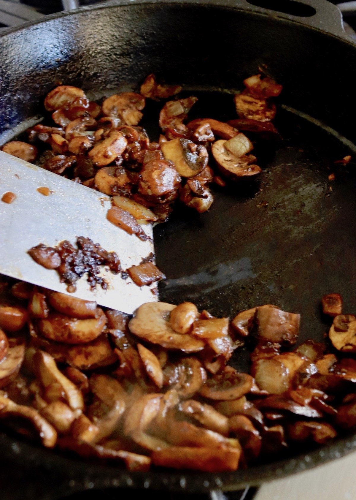 Mushrooms and onion with a metal spatula in cast iron skillet.