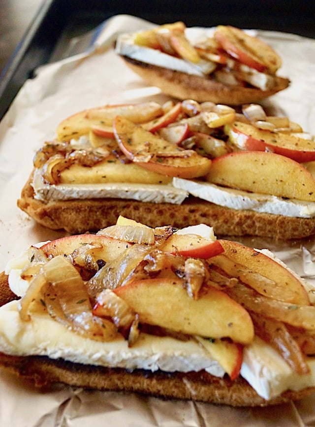 three slices of toast on foil-lined baking sheet, each topped with brie, and cooked onion-apple mixture