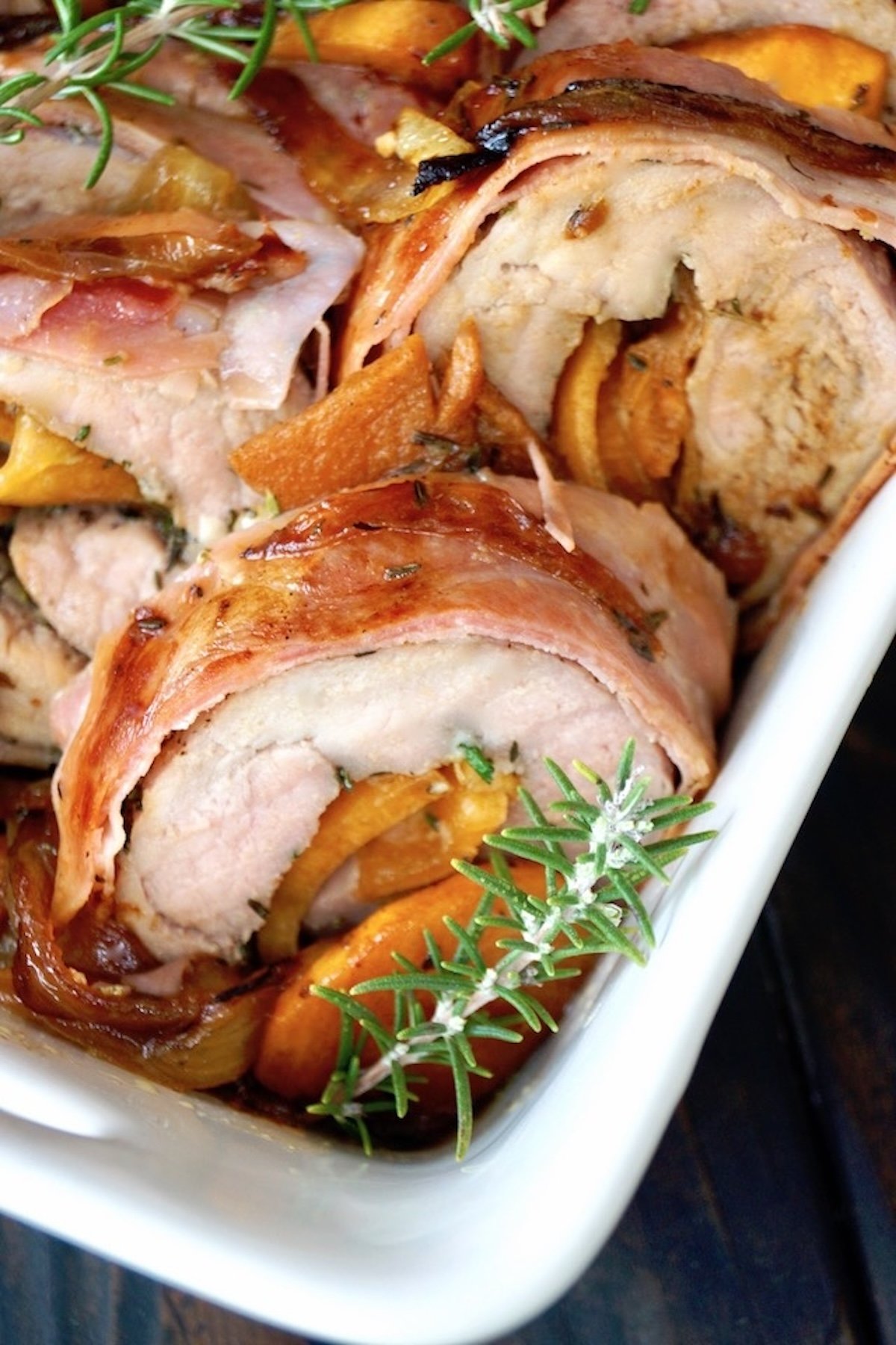 Close up of Persimmon Prosciutto Pork Tenderloin sliced in a white dish with rosemary sprigs.