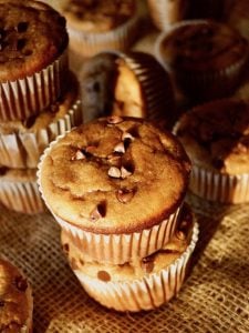 Stack of 2, and stack of 3 gluten-free peanut butter banana muffins with mini chocolate chips, with a few more muffins int he background, on burlap, in the sunlight.