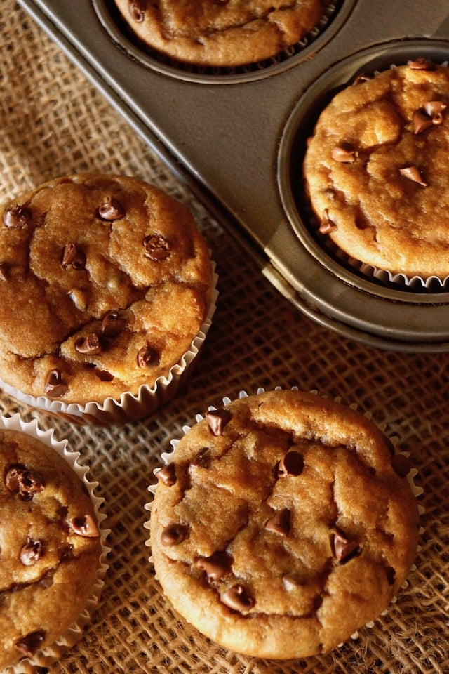 3 gluten-free peanut butter banana muffins with mini chocolate chips on burlap, next to muffin tin with muffins in it.