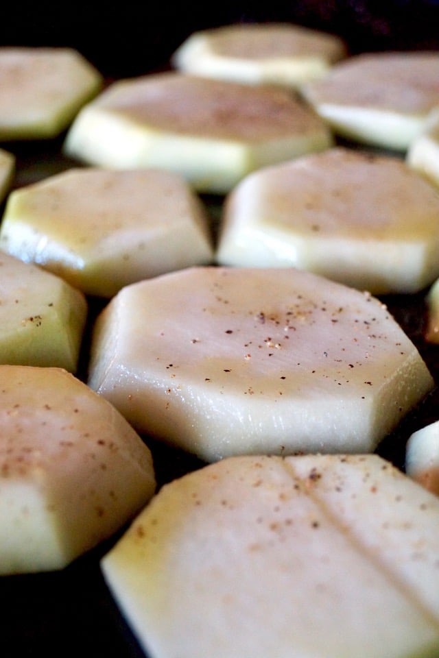 Several slices of raw kohlrabi on a baking sheet with salt, pepper and olive oil, for Roasted Kohlrabi