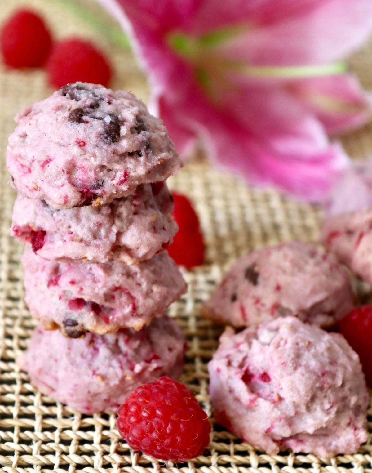 Stack of 4 raspberry chocolate chip cookies with a pink lily flower behind them, with a few fresh bright red raspberries around them.